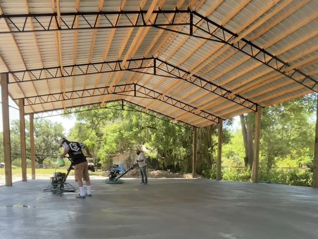 Concrete finisher running a power float across a fresh slab under a pole-barn roof